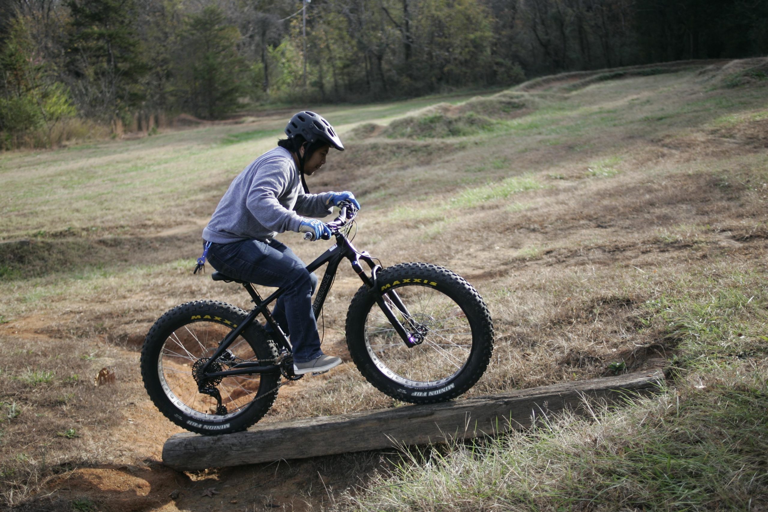 A person wearing a helmet and gloves is riding a black fat bike over a wooden log in a grassy area with trees in the background. The rider is focused on navigating the obstacle, demonstrating an active outdoor biking scene. Hobby Park mountain bike trail.