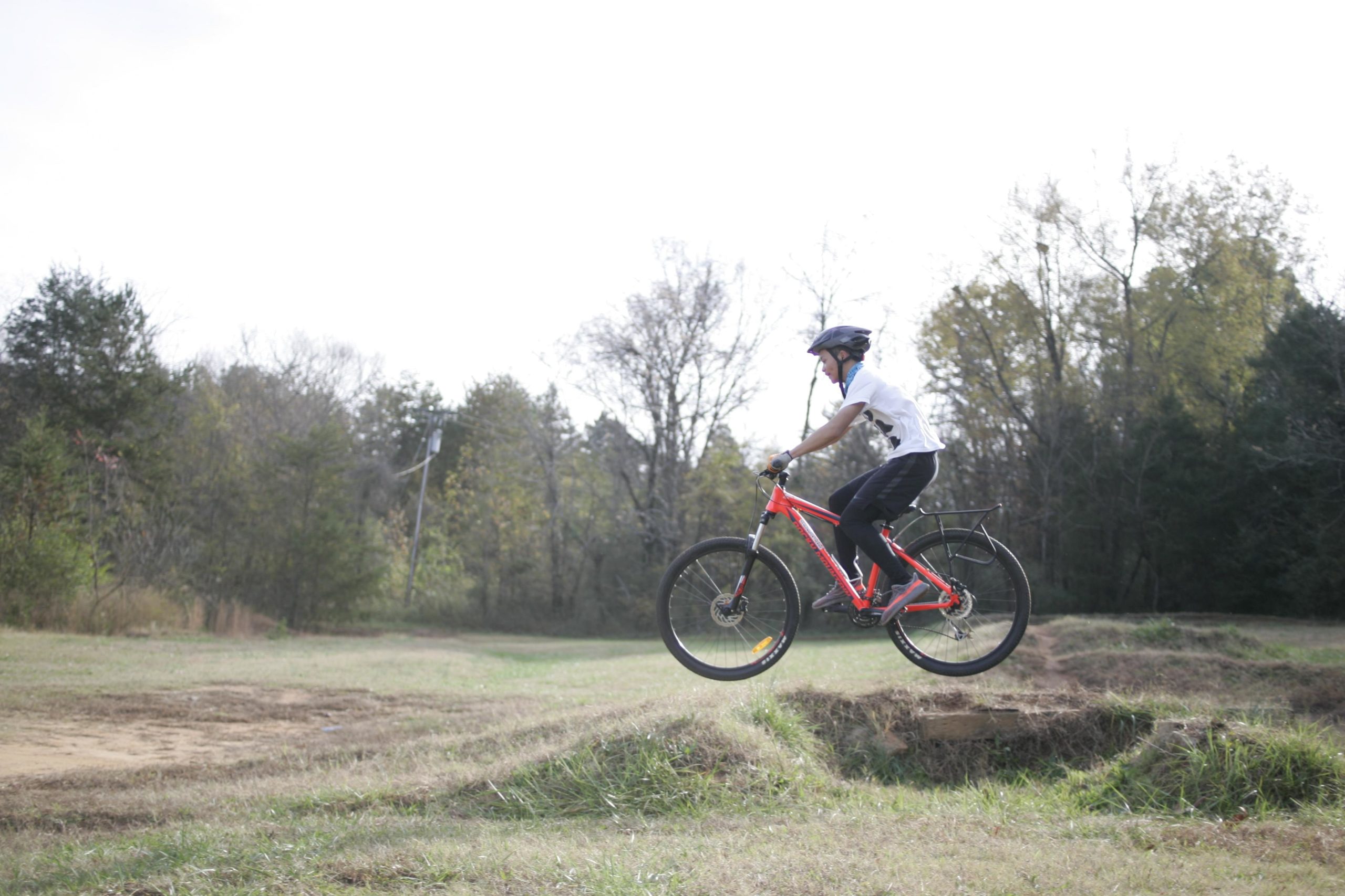 A person in a white shirt and black shorts riding a red mountain bike is mid-air as they jump over a dirt ramp in a grassy field. They are wearing a helmet and appear focused on their landing. Surrounding the area are trees and a clear sky. Hobby Park mountain bike trail.