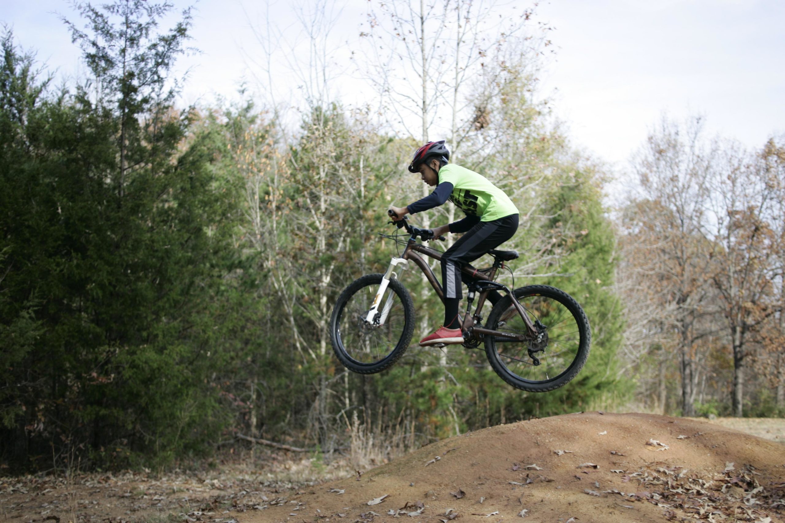 A child in a lime green shirt and black pants is jumping off a dirt ramp on a mountain bike, captured mid-air against a backdrop of trees during a cloudy day. Hobby Park mountain bike trail.