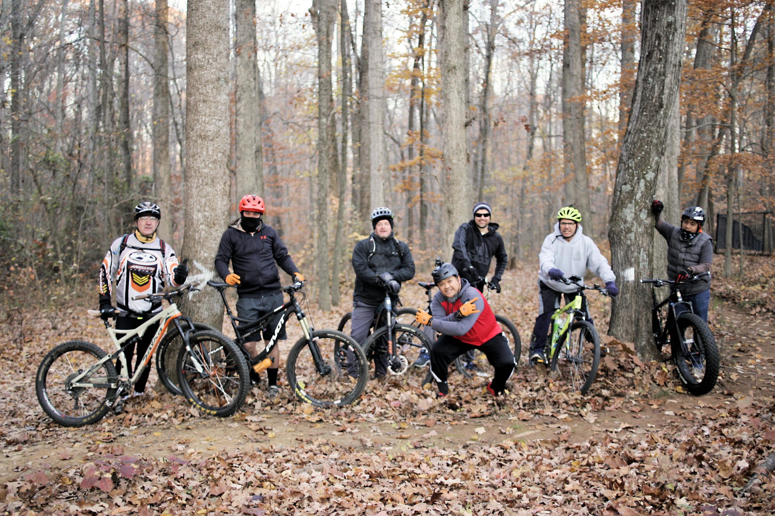 A group of eight people in a forested area during fall, standing next to their mountain bikes. They are wearing helmets and cycling gear, with some individuals posing for the camera. The ground is covered with fallen leaves, and trees with autumn foliage surround them. Deadfall Trail mountain bike trail.