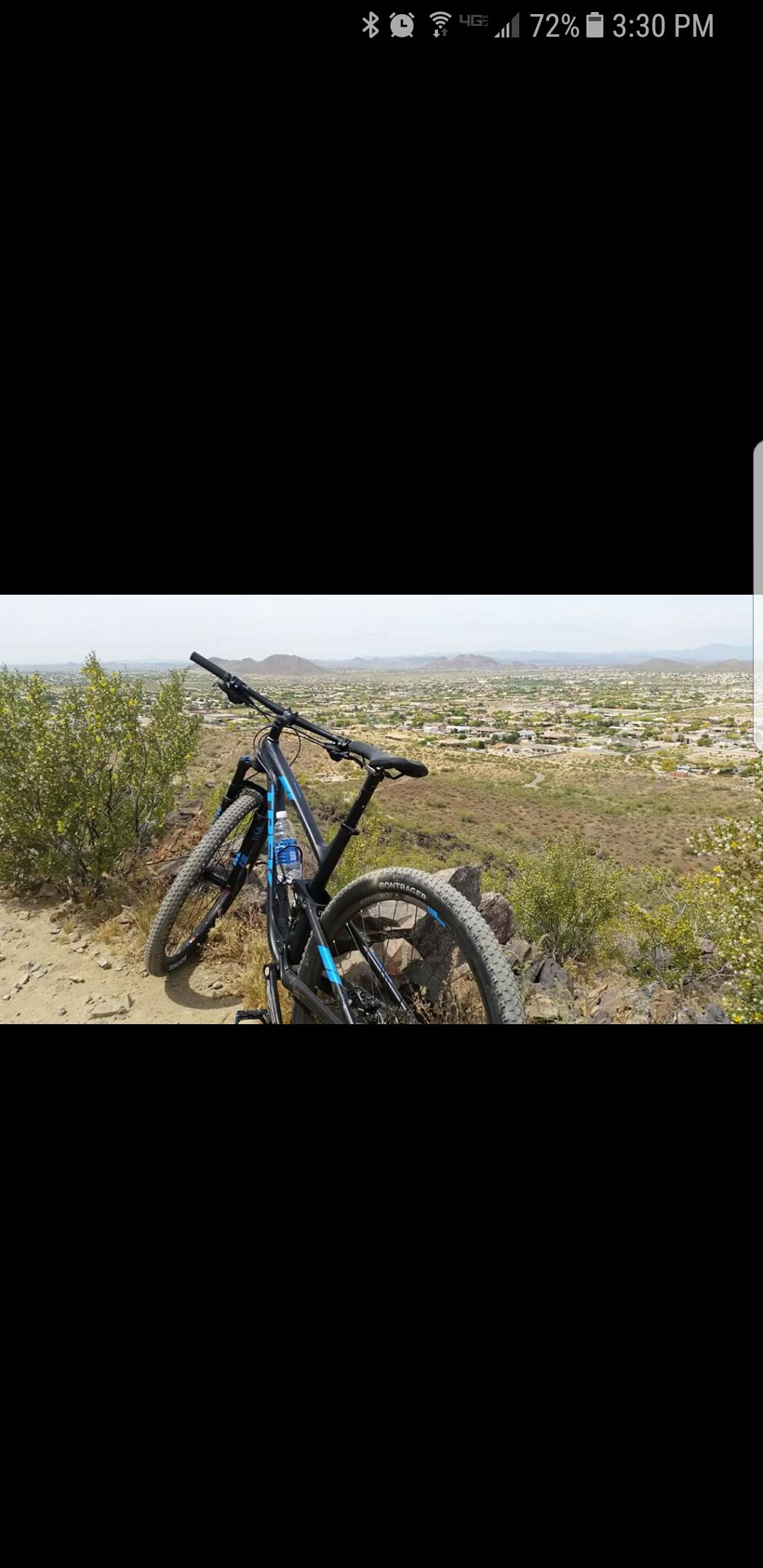 Trek Top Fuel 8: A mountain bike rests on a rocky trail overlooking a vast desert landscape, featuring rolling hills and a distant view of a town below. Bright green bushes are visible alongside the trail, and the sky is partially cloudy.