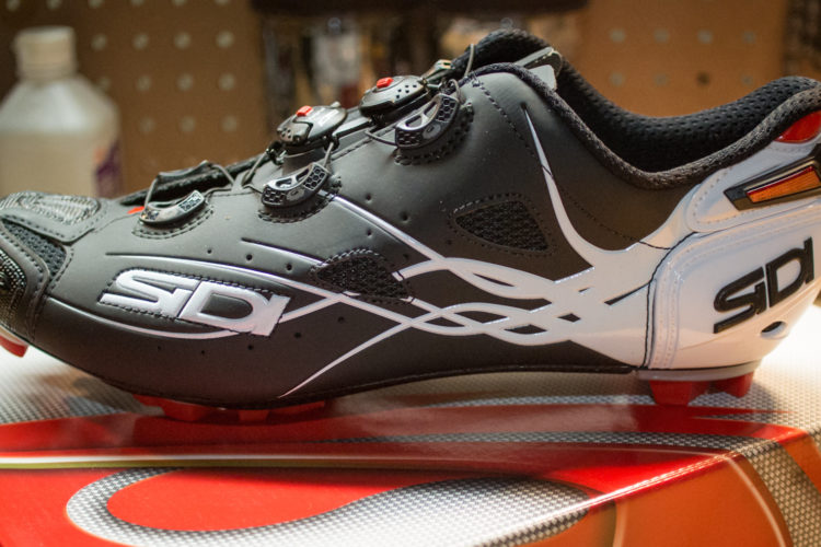 A black and white cycling shoe with red accents, featuring a sleek design and multiple adjustment dials on the upper. The shoe is positioned on a red and gray box with a textured surface, against a backdrop of a pegboard displaying various tools and bottles.