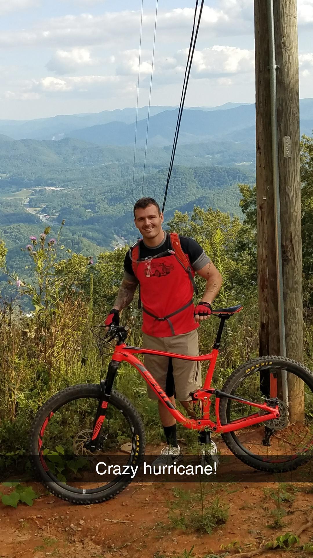 A person is standing next to a bright red mountain bike on a hillside, with a scenic view of mountains and clouds in the background. The individual is wearing a red vest, gloves, and shorts, smiling at the camera. There are power lines and some vegetation around, along with a caption at the bottom reading "Crazy hurricane!" Fire Mountain Trail System mountain bike trail.