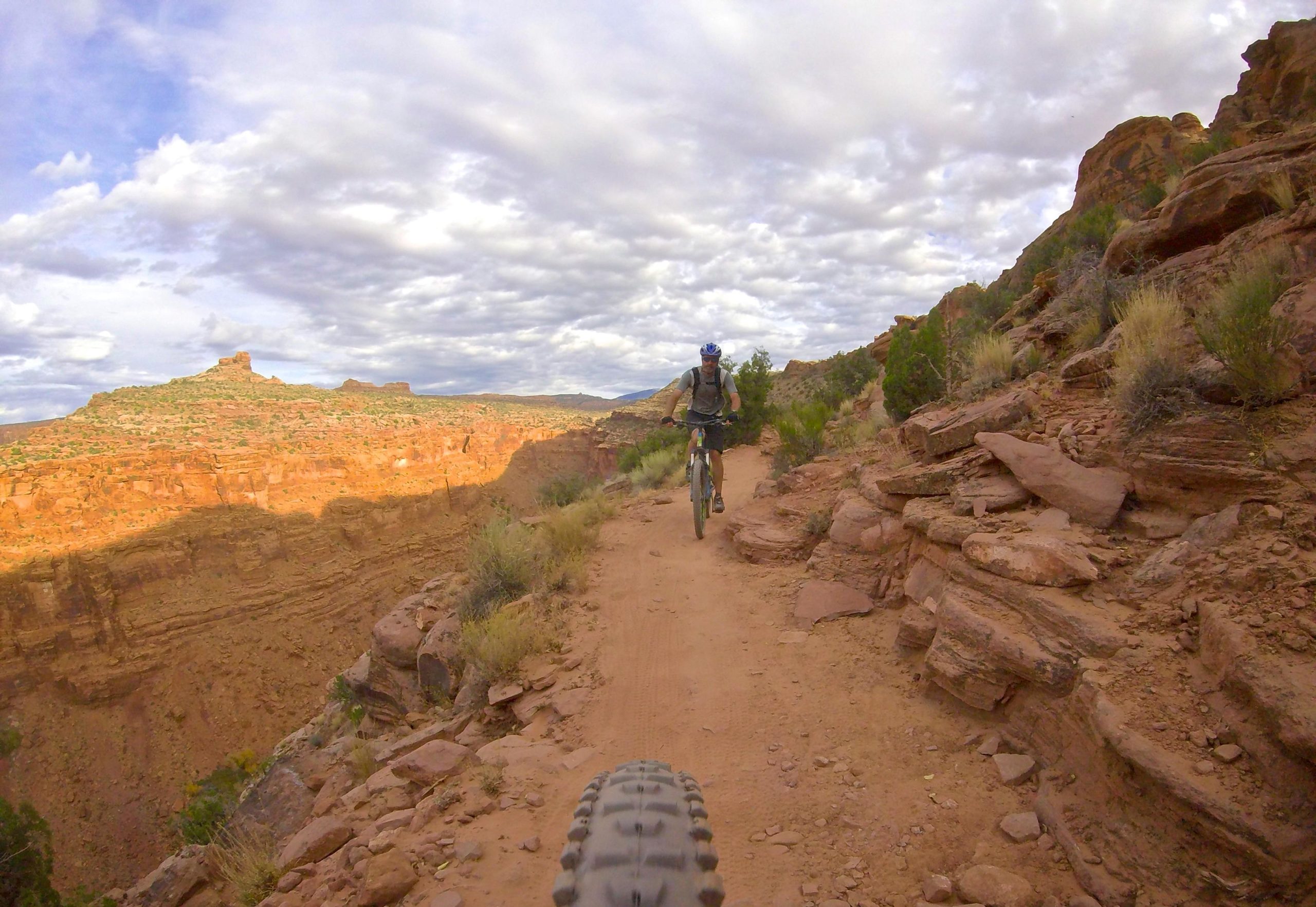 A cyclist riding on a dirt trail surrounded by rocky terrain and shrubs, with a scenic view of a canyon in the background and cloudy skies overhead. The image captures the essence of outdoor mountain biking in a rugged landscape. The Whole Enchilada mountain bike trail.