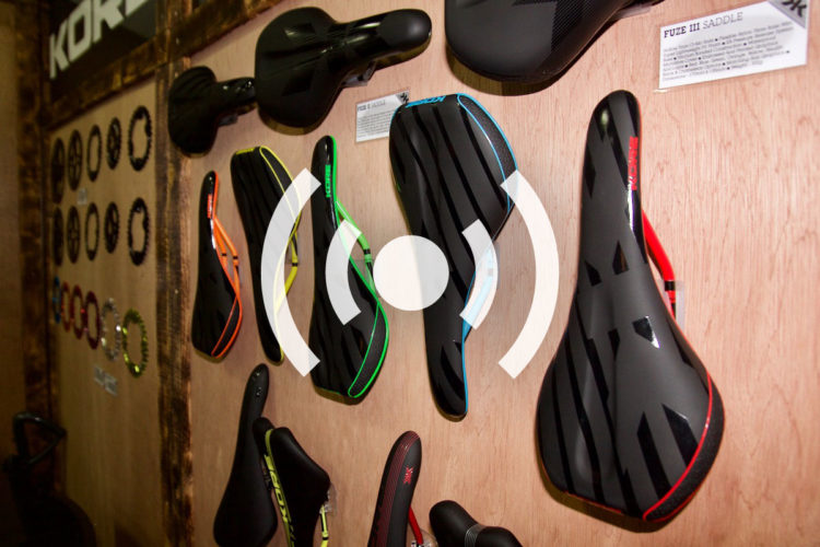 "Display of various bike saddles hanging on a wooden wall, featuring sleek designs in black with colorful accents. Each saddle is labeled for identification, showcasing a range of shapes and styles for cycling enthusiasts."