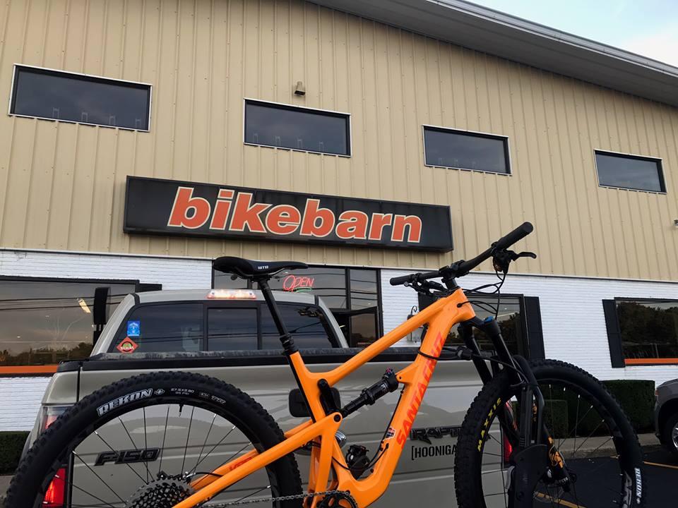 A close-up view of a bright orange mountain bike parked in front of a building with the sign "bikebarn" above its entrance. A pickup truck is visible in the background, and the shop displays an "Open" sign in the window. The building has a light-colored exterior and large windows.