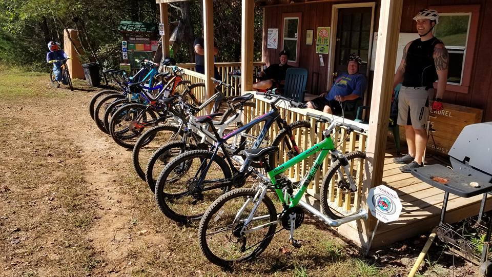A group of mountain bikers relaxing at a trailhead. Several bicycles are lined up along a porch, with two people seated and one standing nearby. The scene is set in a natural outdoor area, showcasing a sunny day with trees in the background. A trail conditions sign is visible, indicating the location is for mountain biking. Mountain Laurel Trails mountain bike trail.