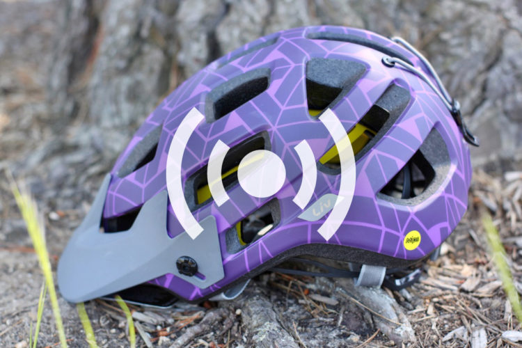 A purple cycling helmet with a geometric pattern resting on the ground, featuring ventilation holes, a gray visor, and a yellow MIPS logo, set against a natural background of tree bark and foliage.