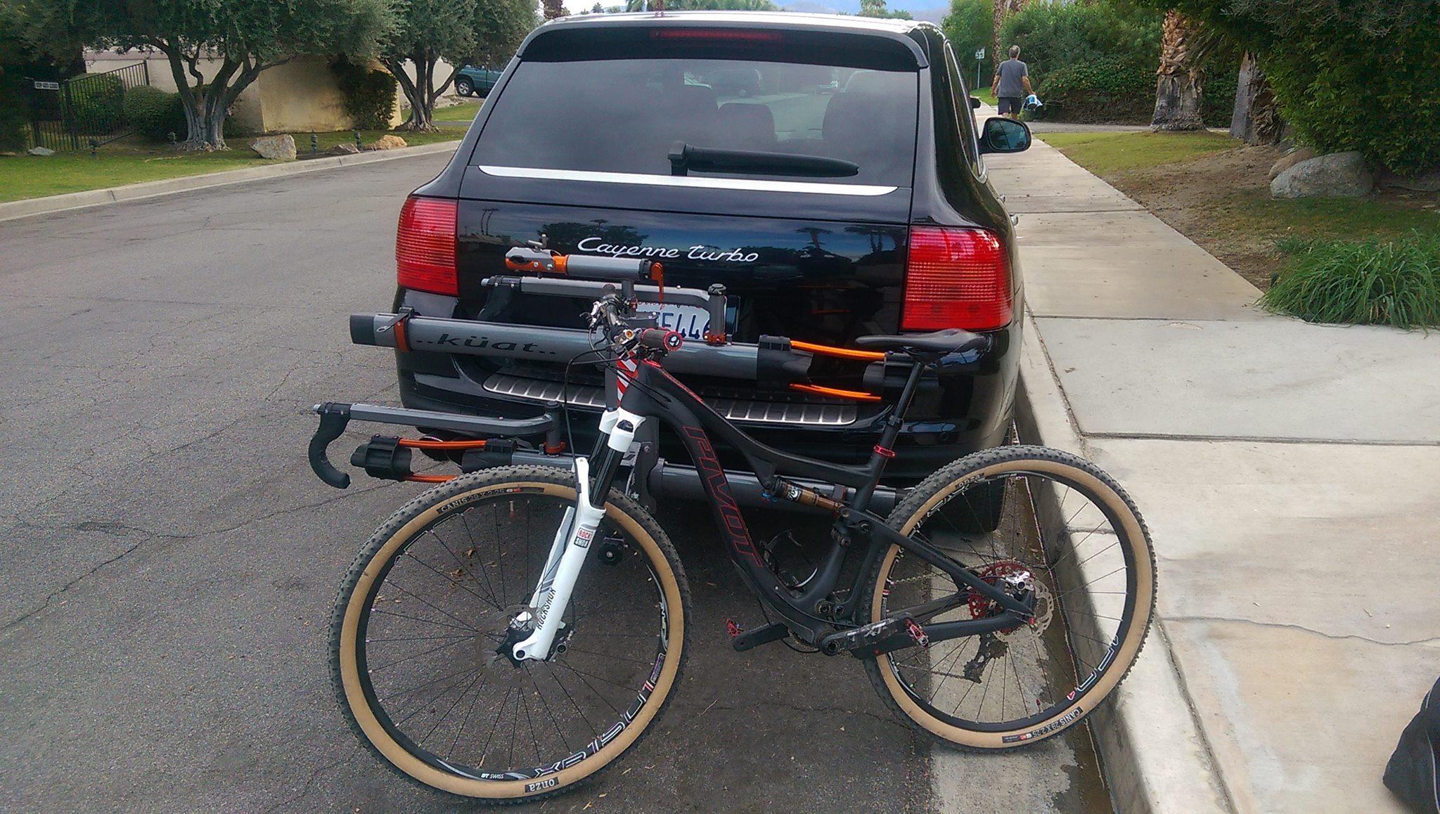 Pivot Mach 429SL Carbon: A black SUV parked on the side of a road with a bicycle secured on a bike rack mounted at the back. The bike features a distinctive frame with a combination of black and white colors, and it has knobby tires suited for off-road riding. In the background, a person walks on the sidewalk, and there are trees and grass lining the street.