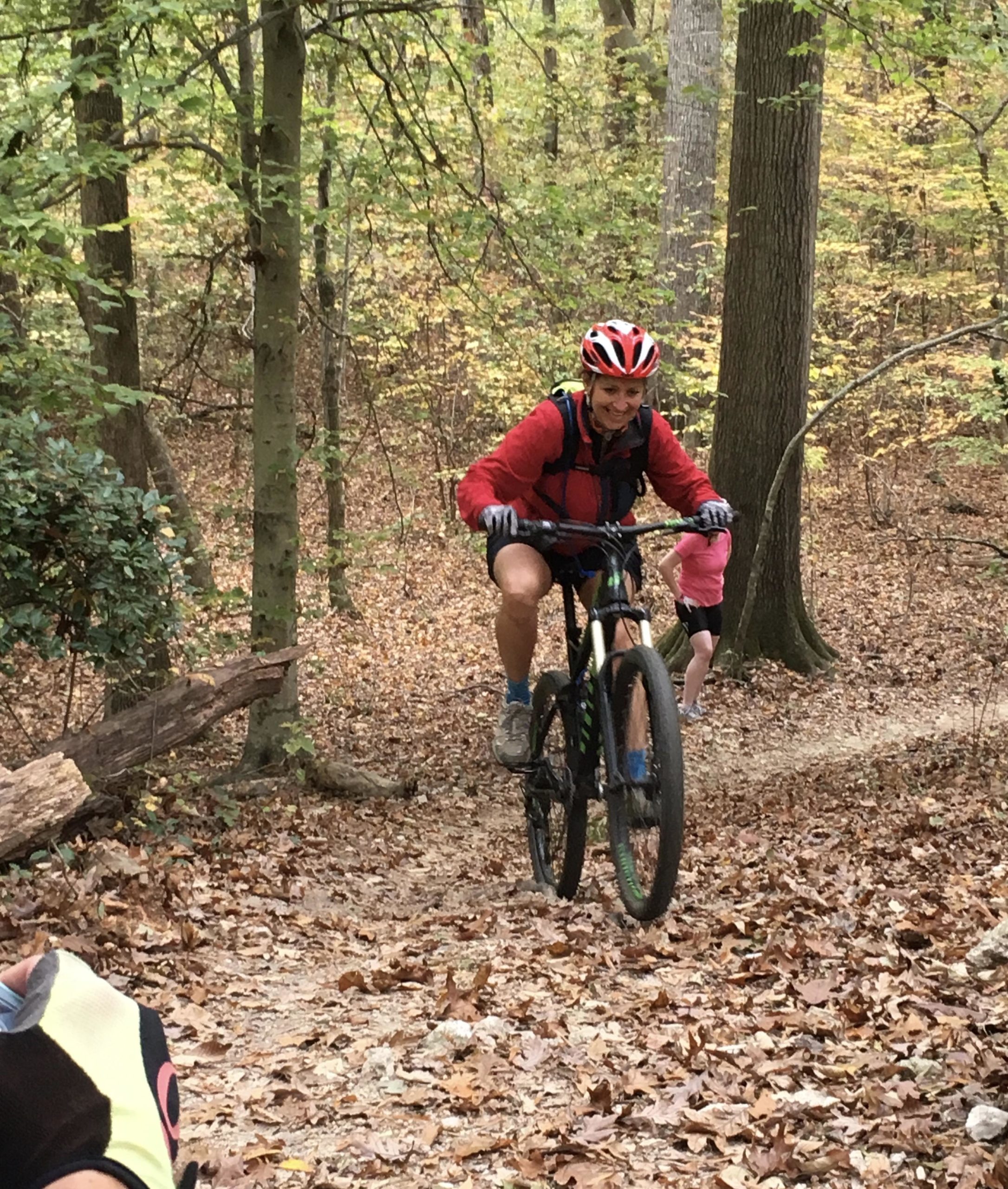 A person riding a mountain bike on a dirt trail in a wooded area, surrounded by autumn leaves and trees. The cyclist is wearing a red jacket and a helmet, smiling as they navigate the trail, while another person in the background appears to be walking. Tanglewood Park mountain bike trail.