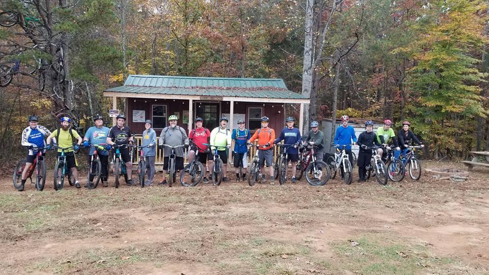 A group of cyclists poses with their mountain bikes outside a wooden building in a forested area. The scene is set in autumn, with trees showcasing colorful leaves. The group consists of both men and women, wearing helmets and cycling gear, ready for a biking adventure. Mountain Laurel Trails mountain bike trail.