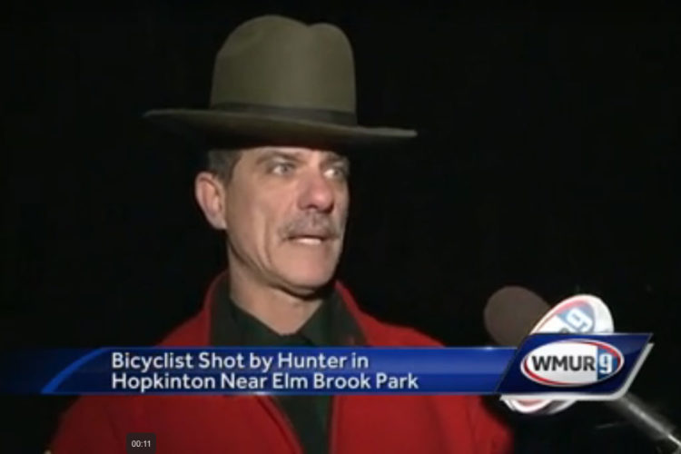 A man wearing a wide-brimmed hat and a red jacket speaks into a microphone during a news segment about an incident involving a bicyclist shot by a hunter in Hopkinton, near Elm Brook Park.