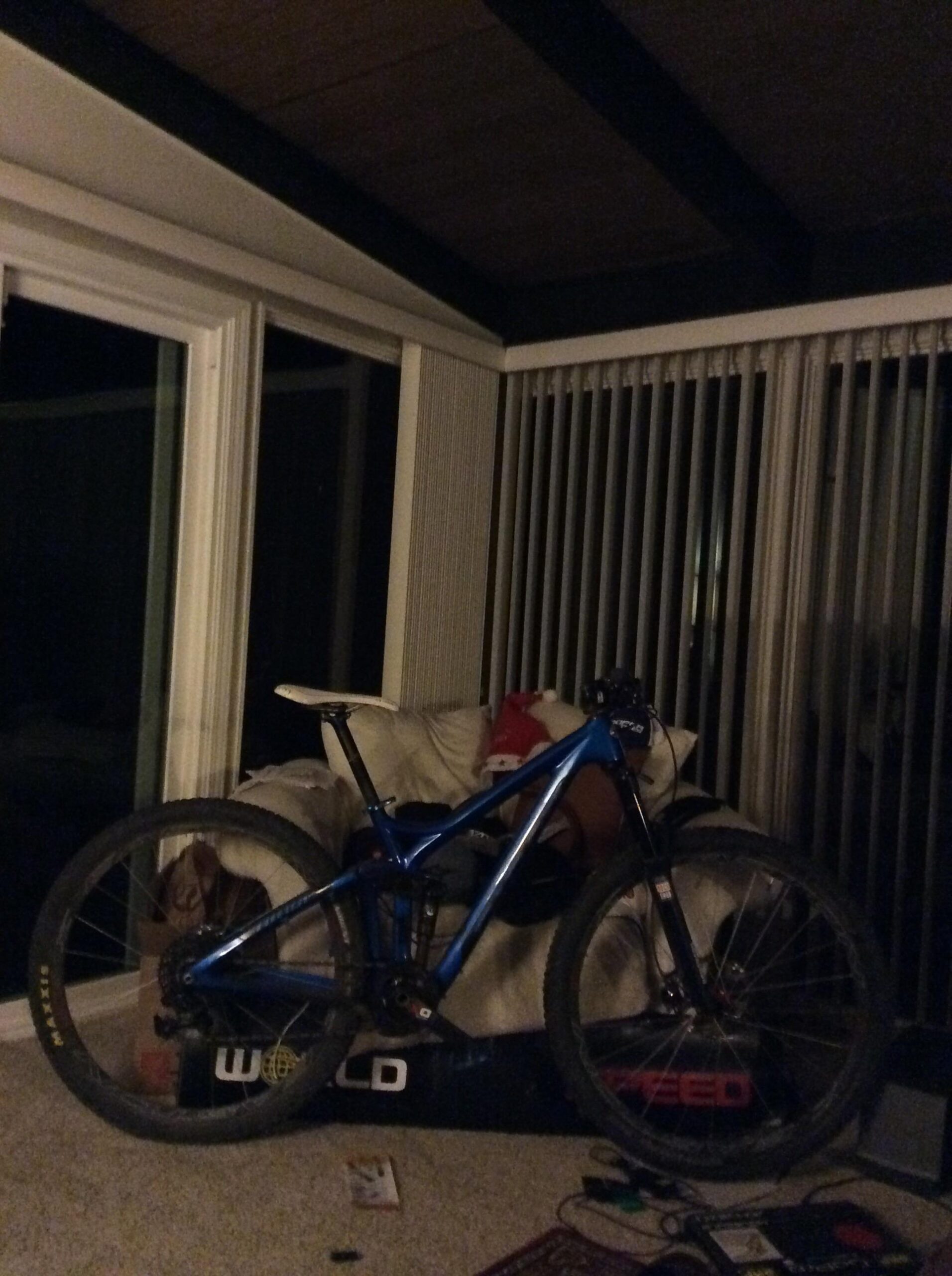 Niner RIP 9: A blue mountain bike leaning against a white couch in a dimly lit room, with large windows and vertical blinds in the background. A small red object is visible on the couch, and various items are scattered on the floor.
