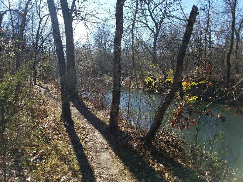 A serene, sunlit path along a river, bordered by tall trees and sparse underbrush. The water reflects the blue sky, and shadows from the trees stretch across the trail, enhancing the peaceful nature scene. Sac River Trail mountain bike trail.