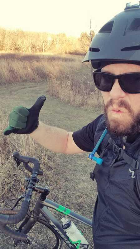 A person wearing a black helmet and sunglasses gives a thumbs-up while standing next to a bicycle in a grassy area. The background features tall grass and trees under a clear sky, suggesting a recreational outdoor setting. Lapham Peak State Park mountain bike trail.