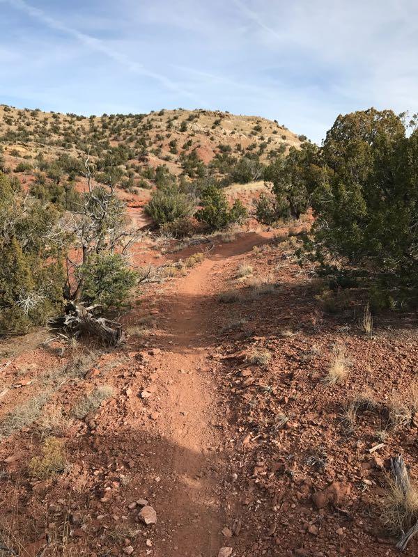 A winding dirt trail cuts through a rocky, reddish landscape, bordered by scattered shrubs and trees. The path leads towards a gently rising hill in the background, under a clear sky with thin clouds. Golden Open Space mountain bike trail.