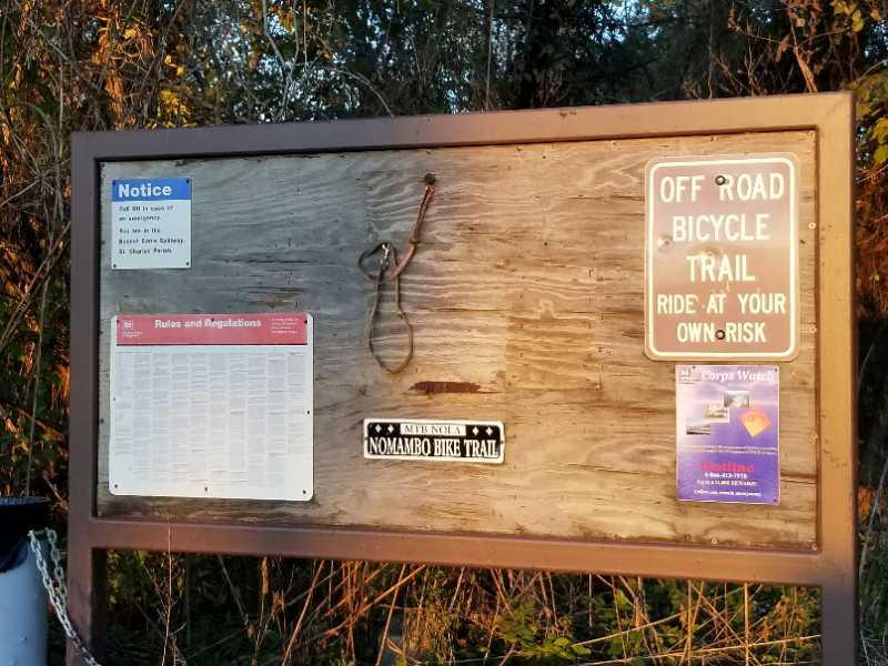 A wooden information board at the entrance of an off-road bicycle trail. The board features a blue notice sign, a list of rules and regulations, and a warning sign that reads "Off Road Bicycle Trail - Ride at Your Own Risk." A small black and white sign states "Nomambo Bike Trail." The surrounding area is lush with greenery. Bonnet Carre Spillway Trail mountain bike trail.