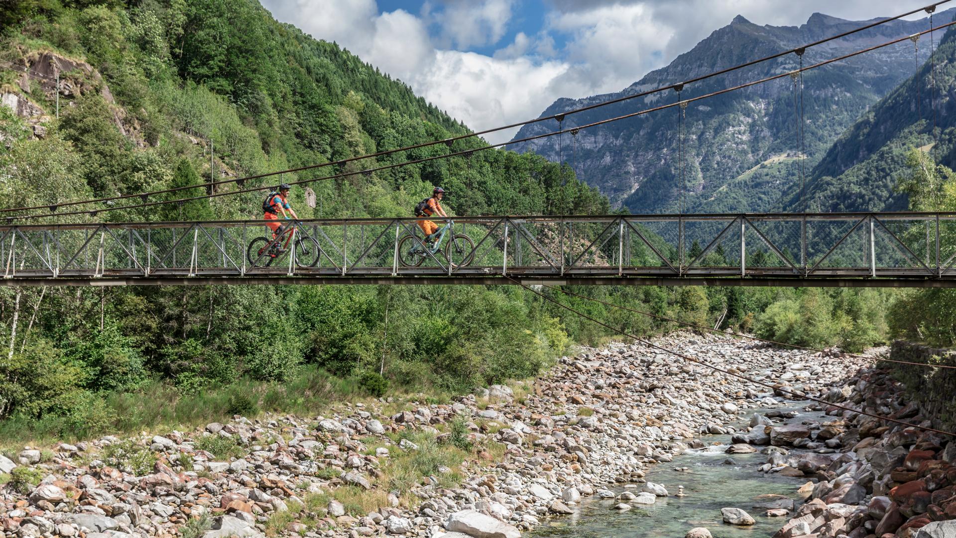 Two mountain bikers ride across a suspension bridge over a rocky riverbed, surrounded by lush greenery and mountains under a partly cloudy sky. Alta Verzasca Bike mountain bike trail.