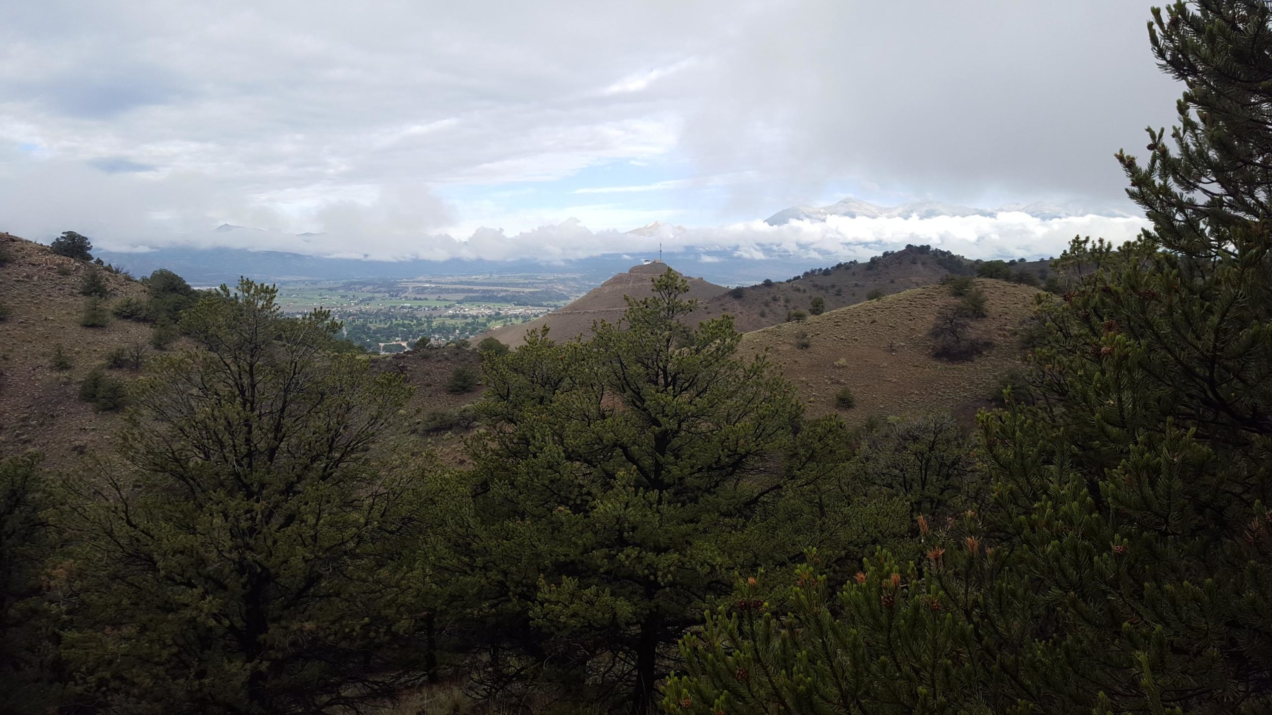 A panoramic view of hills and valleys under a cloudy sky, featuring a mix of greenery and rocky terrain. In the distance, mountains are partially obscured by clouds, creating a serene natural landscape. Arkansas Hills mountain bike trail.