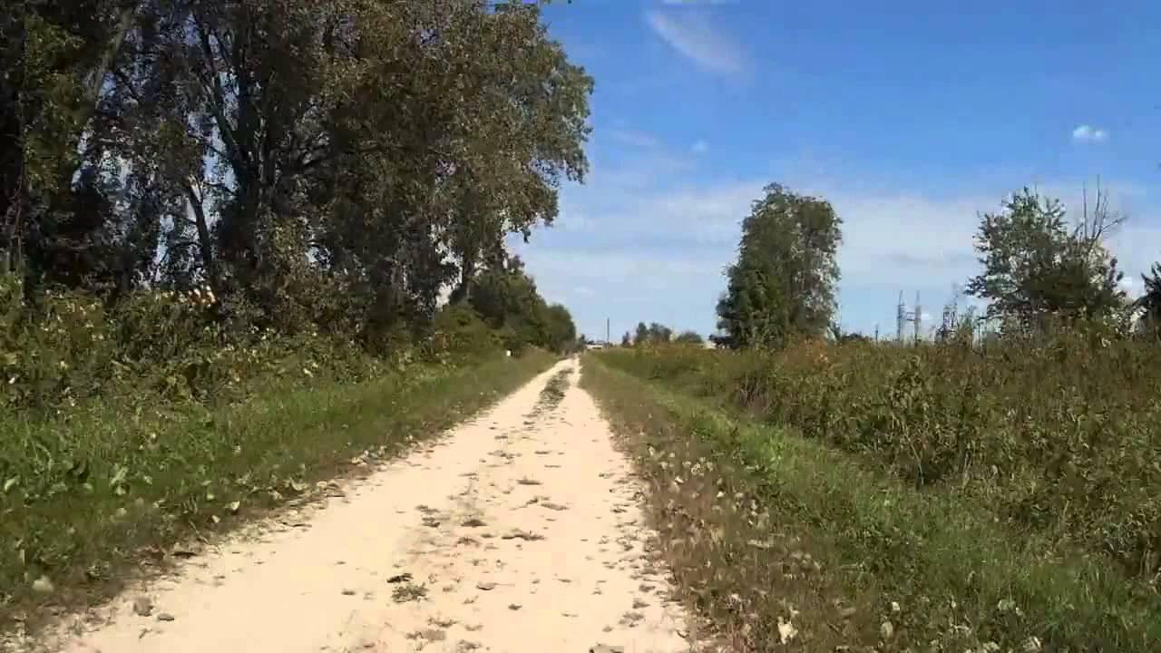 A sandy path lined with greenery, leading through a landscape with trees on both sides. The sky is blue with a few clouds, indicating a sunny day. Wauponsee Glacial Trail mountain bike trail.