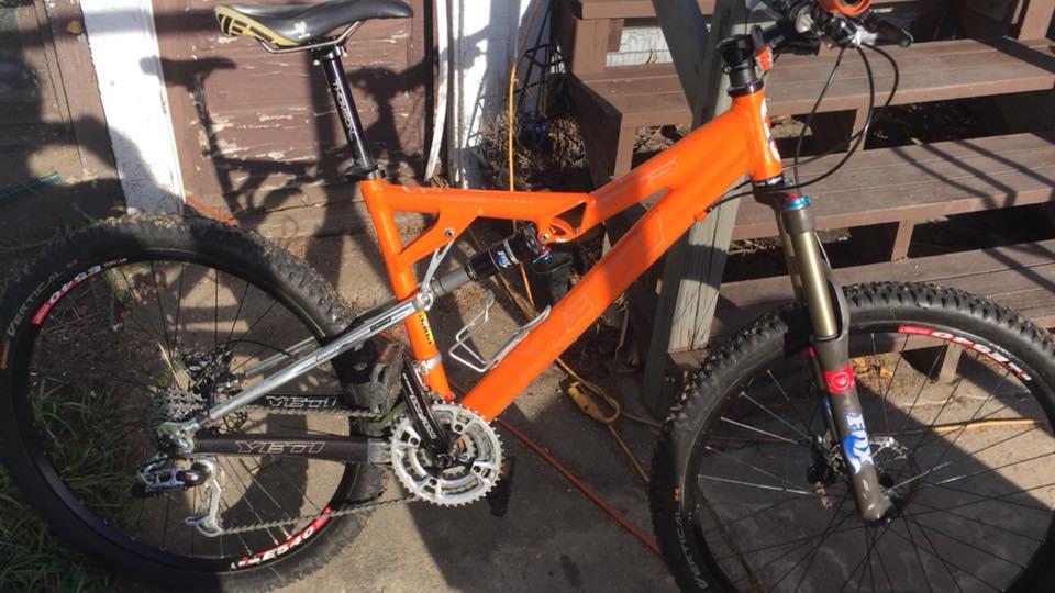 Yeti AS-R (Alloy): A close-up image of an orange mountain bike leaning against a wooden structure, showcasing its components including a suspension fork, chain, and tires. The bike features a sleek design with a prominent brand logo on the frame, and it rests on a pavement area with some grass visible nearby.
