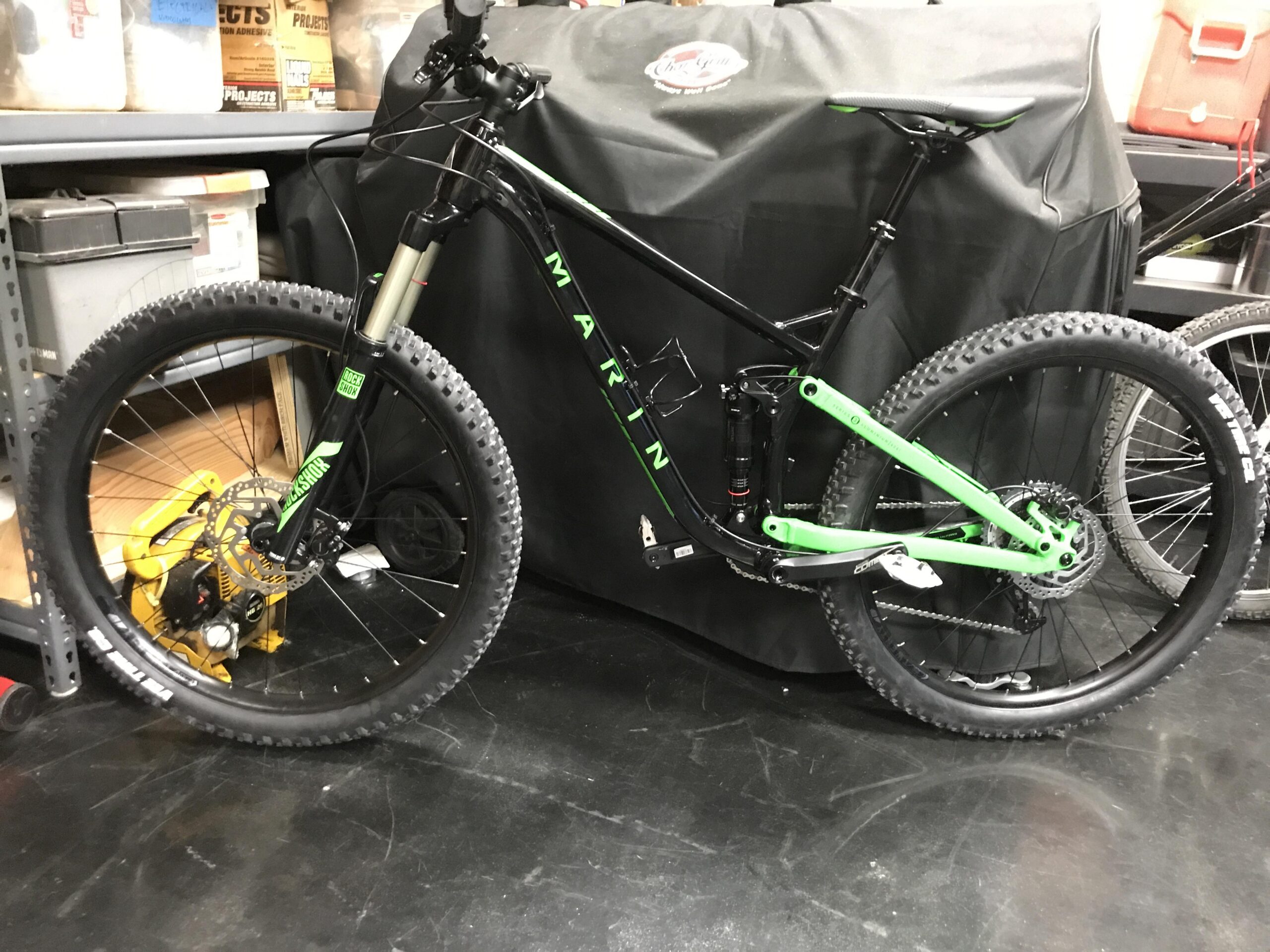 Marin Marin B-17 1: A mountain bike with a black frame and green accents leaned against a black covered object in a garage setting. The bike features thick tires, a front suspension fork, and disc brakes, with various storage containers visible in the background.