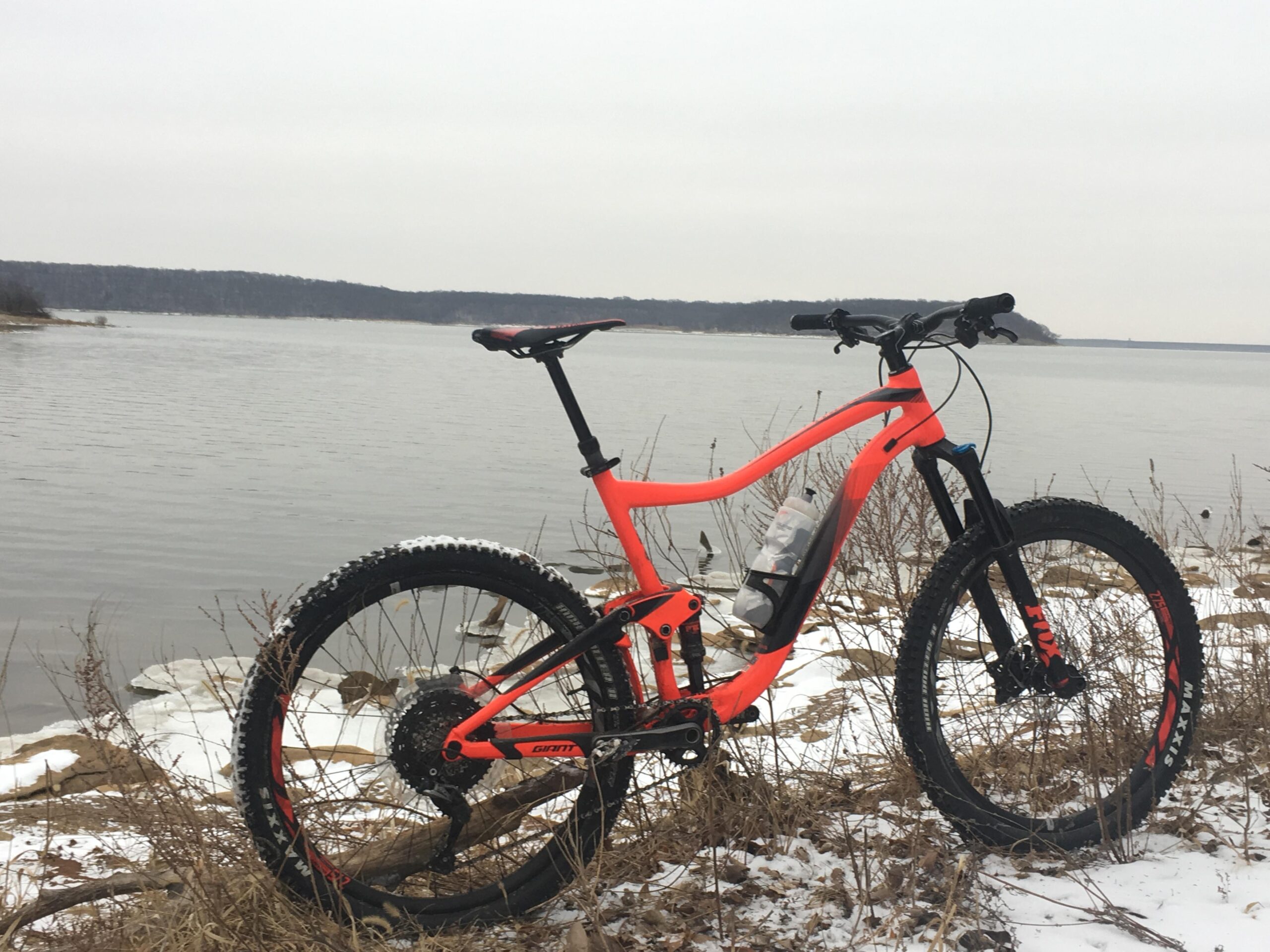 Giant Trance 27.5 2: A bright orange mountain bike is positioned near a snowy shoreline, with a calm lake in the background. The bike's tires are partially covered in snow, and the landscape features sparse vegetation and distant trees. The sky is overcast, creating a muted winter atmosphere.