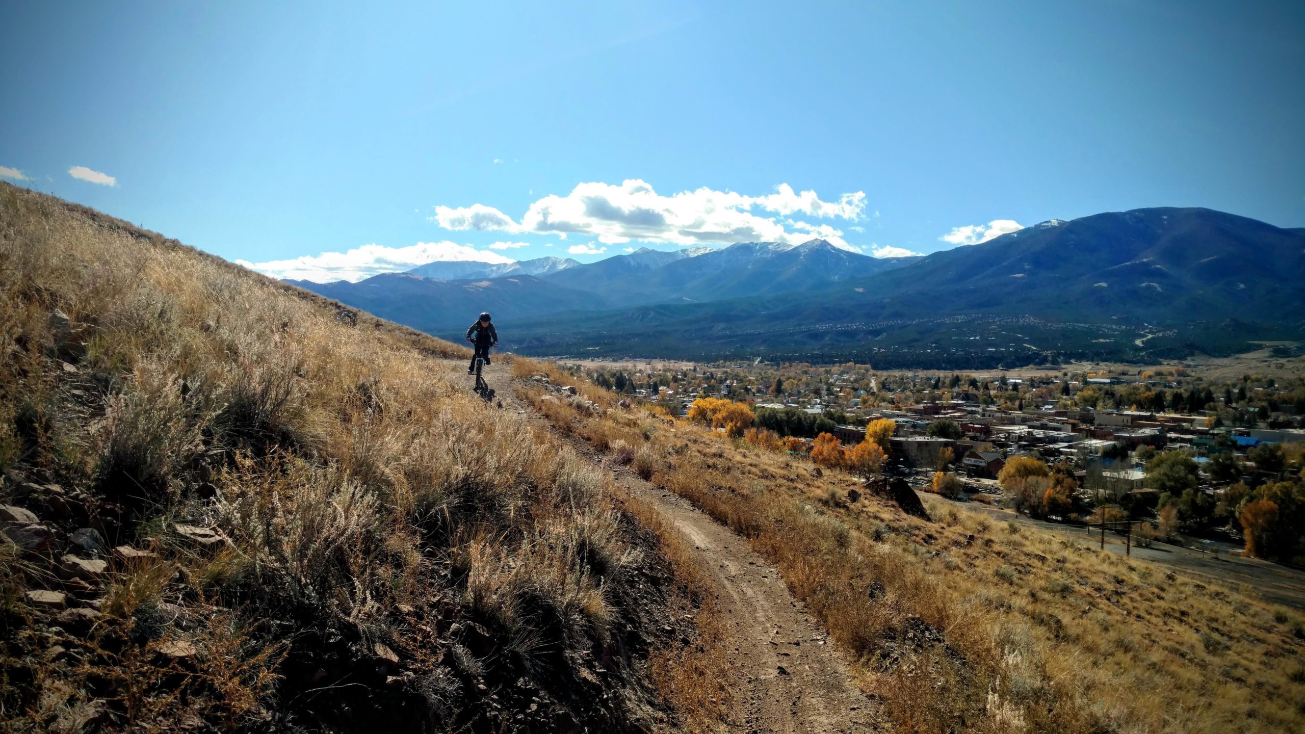 A person riding a mountain bike along a hillside trail, surrounded by tall grass, with a scenic view of a valley and mountains in the background under a clear blue sky. Arkansas Hills mountain bike trail.