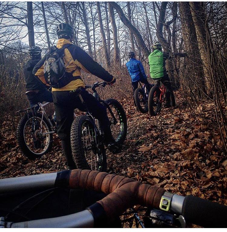 Four cyclists riding fat bikes on a leaf-covered trail in a wooded area. The group can be seen from behind, wearing jackets in various colors, and the handlebars of a bike are in the foreground. The trees in the background are bare, indicating a late autumn or early winter season. Western University trails mountain bike trail.