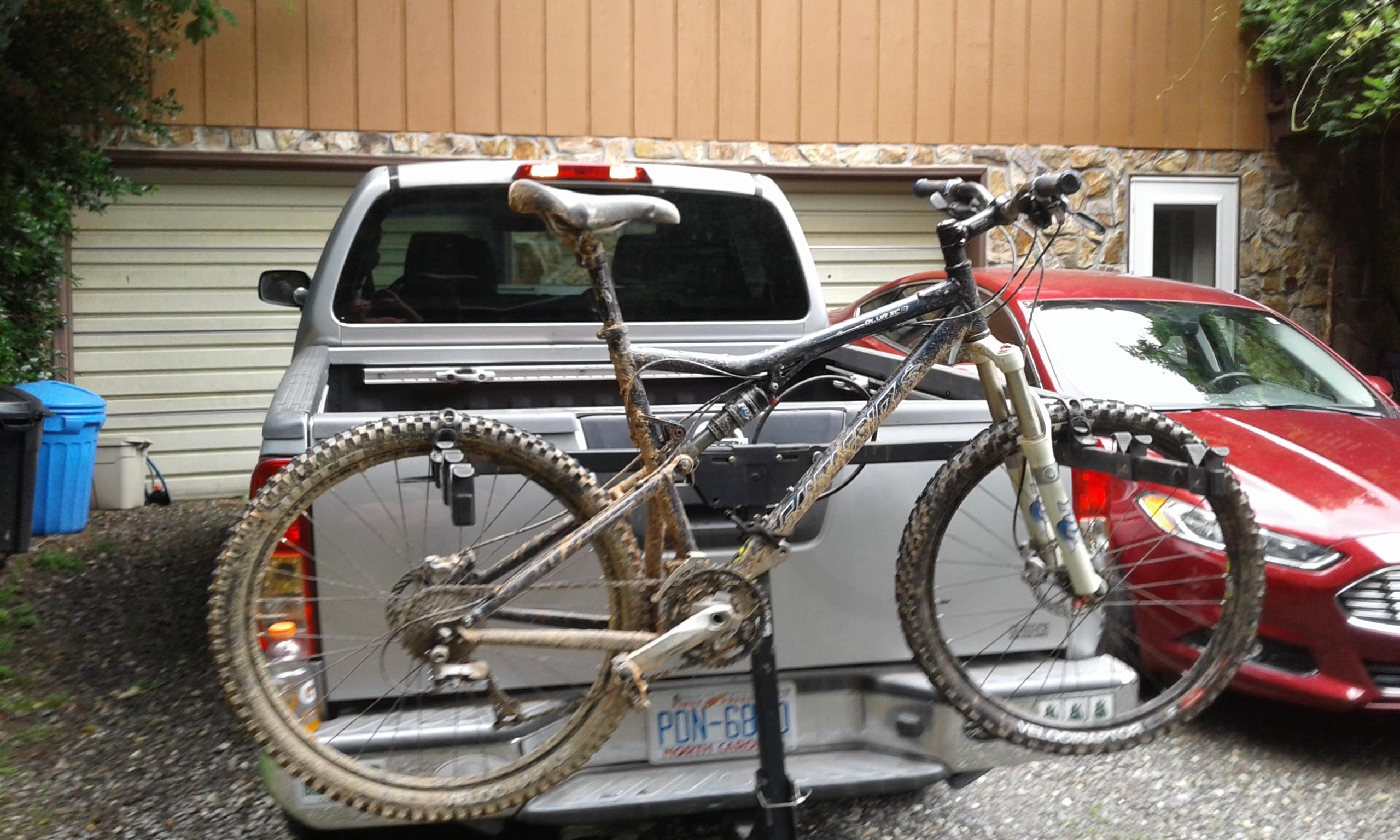 Santa Cruz Blur: A muddy mountain bike mounted on the back of a silver pickup truck, parked in a driveway. A red car is visible in the background, along with a blue trash bin and a garage door. The bike shows signs of recent off-road use, with dirt splattered on the frame and wheels.