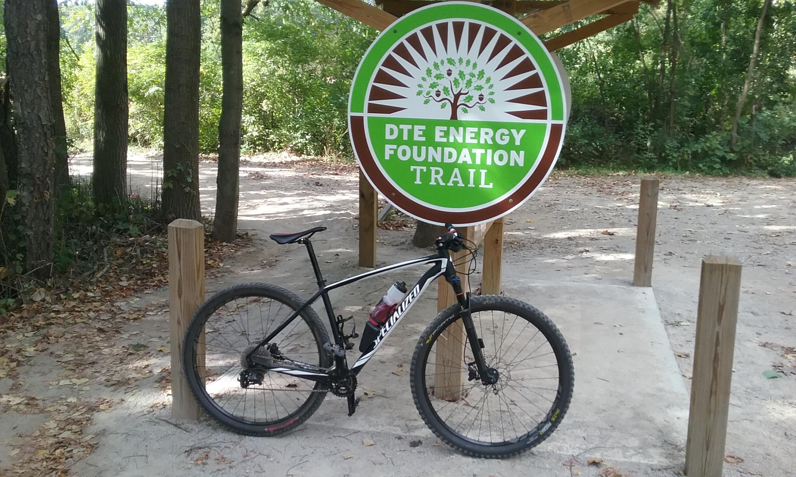 A black mountain bike leaning against a wooden post near a sign for the "DTE Energy Foundation Trail." The sign is prominently displayed, featuring a tree graphic and the trail name, surrounded by wooded scenery and fallen leaves on the ground. DTE Energy Foundation Trail mountain bike trail.