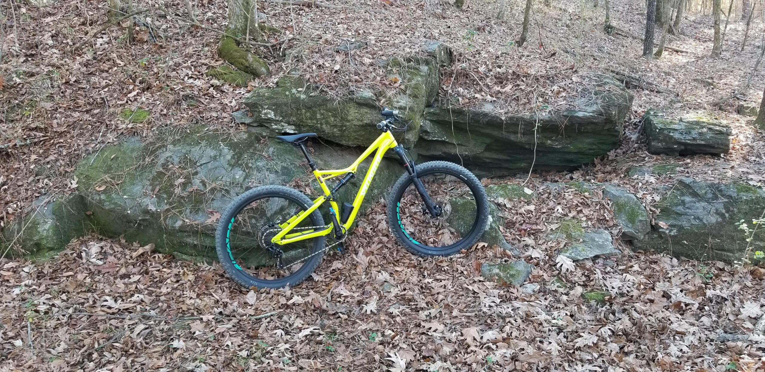 Specialized Stumpjumper FSR Comp 6Fattie: A brightly colored yellow mountain bike is parked next to a large rock in a wooded area covered with fallen leaves. Surrounding trees and more rocks are visible in the background, indicating an outdoor trail setting.