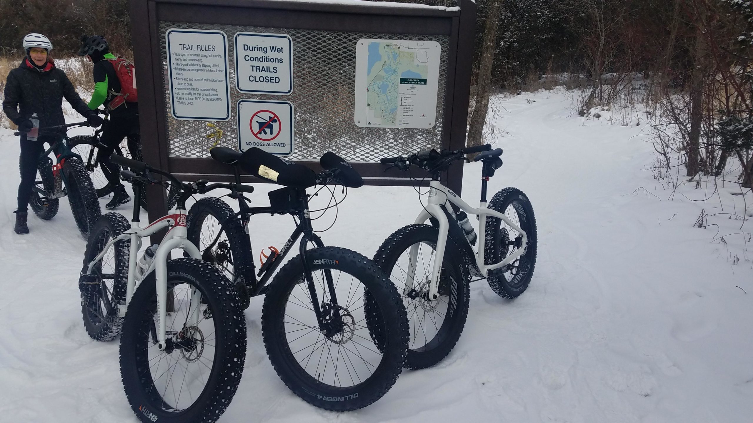A snowy outdoor scene featuring two cyclists standing next to a sign displaying trail rules and a map. In the foreground, three fat-tire bikes are parked in the snow. The background shows a snow-covered trail leading into the woods. Elm Creek Park mountain bike trail.