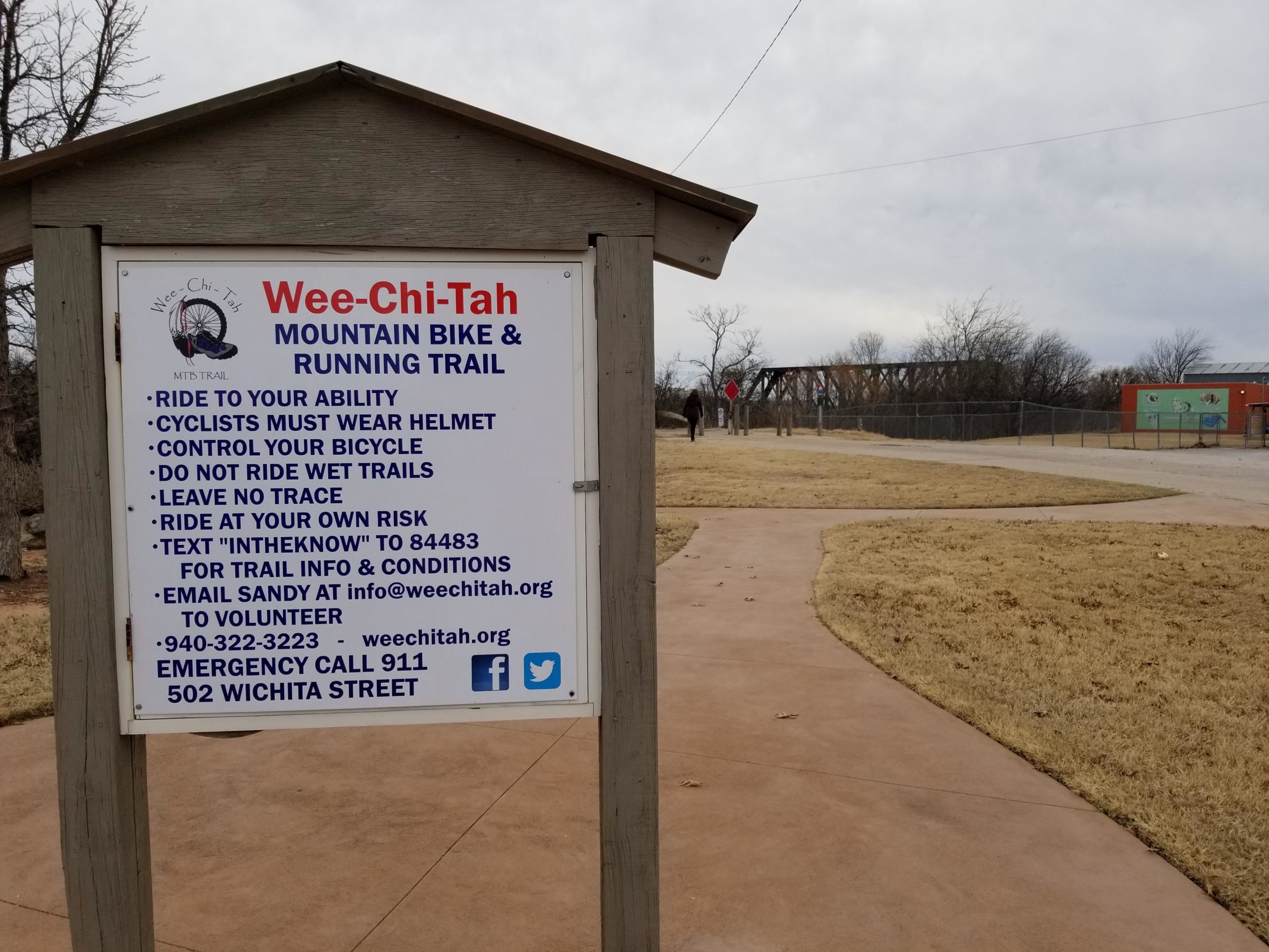 Sign for the Wee-Chi-Tah mountain bike and running trail, featuring safety guidelines and contact information. The sign lists instructions such as wearing a helmet, controlling bicycles, and not riding on wet trails. The surrounding area includes a paved path and a nearby bridge in the background. Wee-chi-ta mountain bike trail.