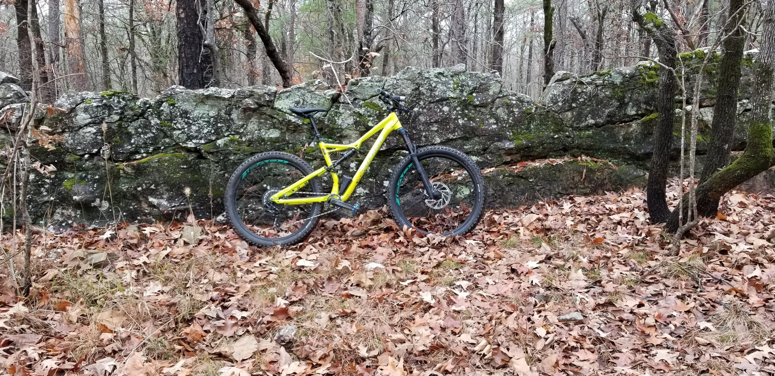 Specialized Stumpjumper FSR Comp 6Fattie: A bright yellow mountain bike leaning against a moss-covered rock wall, surrounded by fallen leaves and sparse vegetation in a wooded area. Trees are visible in the background, indicating a natural outdoor setting.
