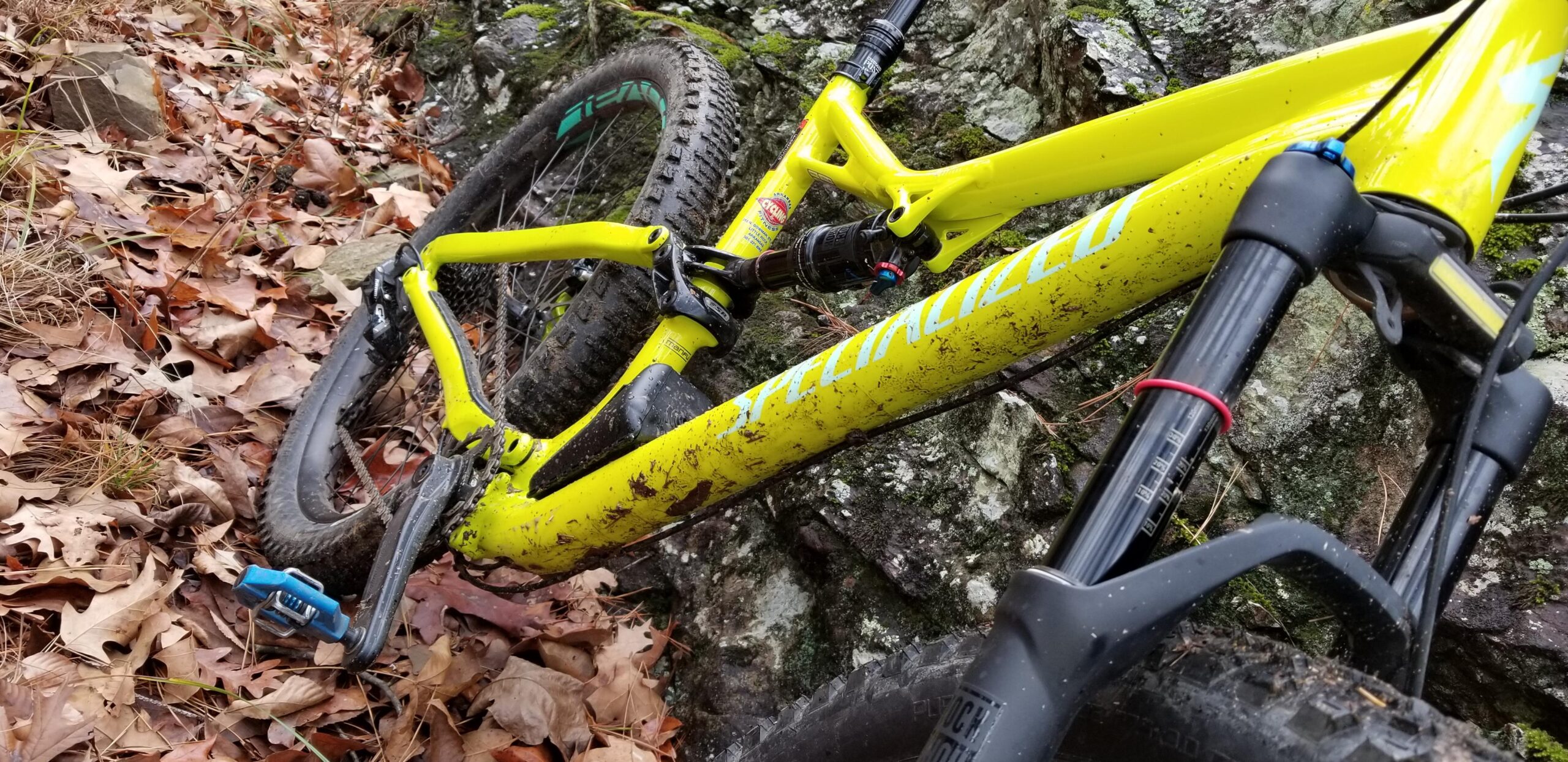 Specialized Stumpjumper FSR Comp 6Fattie: A close-up of a muddy yellow mountain bike leaning against a rock, surrounded by fallen leaves and grass. The bike features a sturdy frame, thick tires, and visible dirt on its components, indicating recent off-road use.