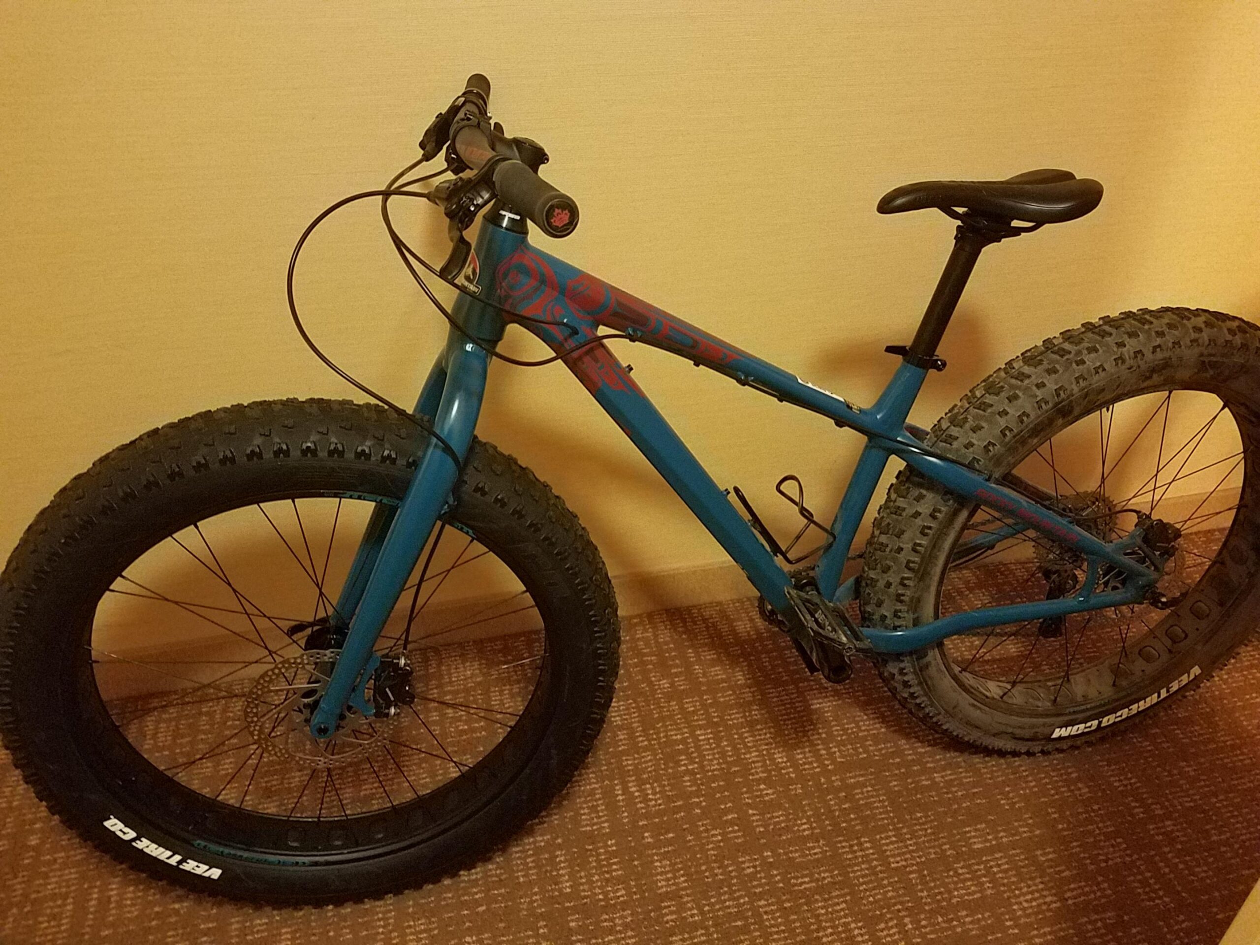 Rocky Mountain Blizzard: A blue fat bike with wide tires, leaning against a wall in an indoor setting. The bike features a black seat, handlebars with grips, and disc brakes on both wheels. The background shows a beige wall and a brown patterned carpet.