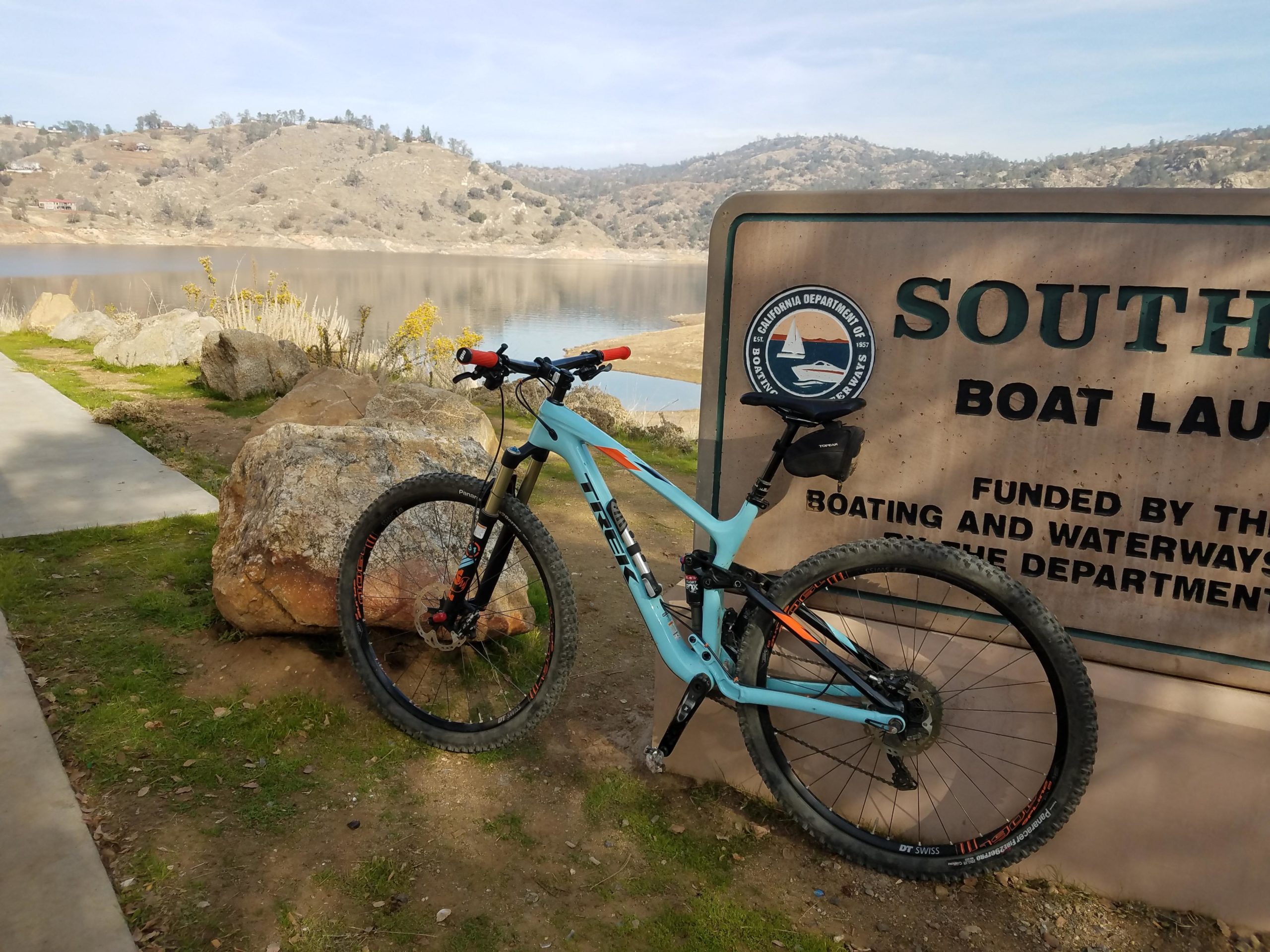 A mountain bike leaning against a sign that reads "South Boat Launch" near a serene lake. The surrounding landscape features rolling hills and bare trees, with rocks and grass along the path. The sky is partly cloudy, providing a peaceful outdoor atmosphere. San Joaquin River Trail mountain bike trail.