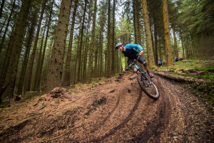 A mountain biker navigating a winding dirt trail through a dense forest, surrounded by tall trees and pine needles on the ground. The rider leans into a turn, with visible motion and determination, while other bikers can be seen in the background.