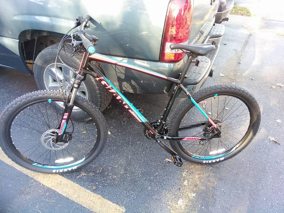 2017 Giant Talon 3: A black mountain bike with colorful accents parked next to a silver pickup truck, featuring thick tires and disc brakes. The bike is positioned on a paved surface with some fallen leaves visible nearby.