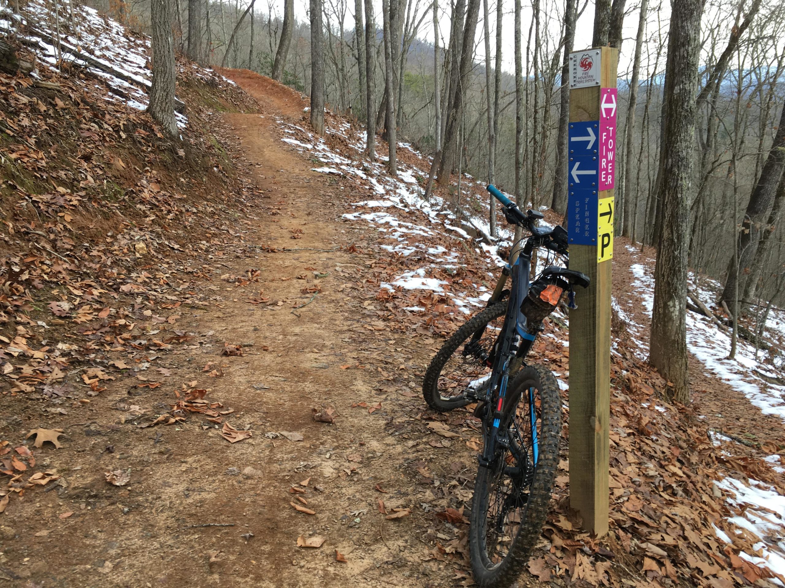 A mountain bike rests against a trail sign on a dirt path surrounded by trees. The ground is covered with fallen leaves and patches of snow, indicating a winter setting. The sign displays directional arrows and trail names, guiding bikers along the route. Fire Mountain Trail System mountain bike trail.