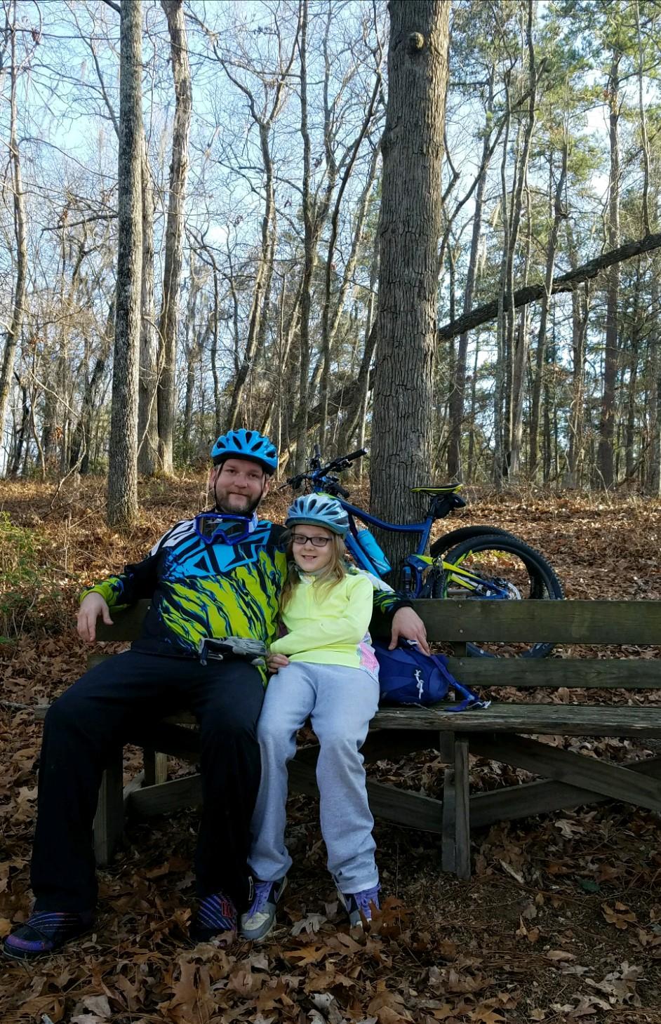 A man and a girl wearing bright safety helmets sit together on a wooden bench in a wooded area. They are smiling, surrounded by trees with bare branches and fallen leaves on the ground. A mountain bike is parked behind them. The Knot mountain bike trail.