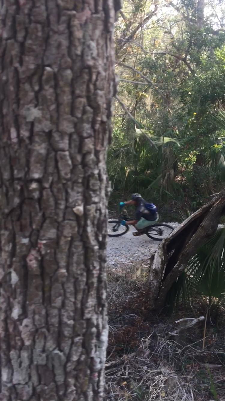 A mountain biker riding along a gravel trail in a dense forest, partially obscured by a tree trunk in the foreground. Lush greenery surrounds the path, creating a natural outdoor setting. Mala Compra mountain bike trail.