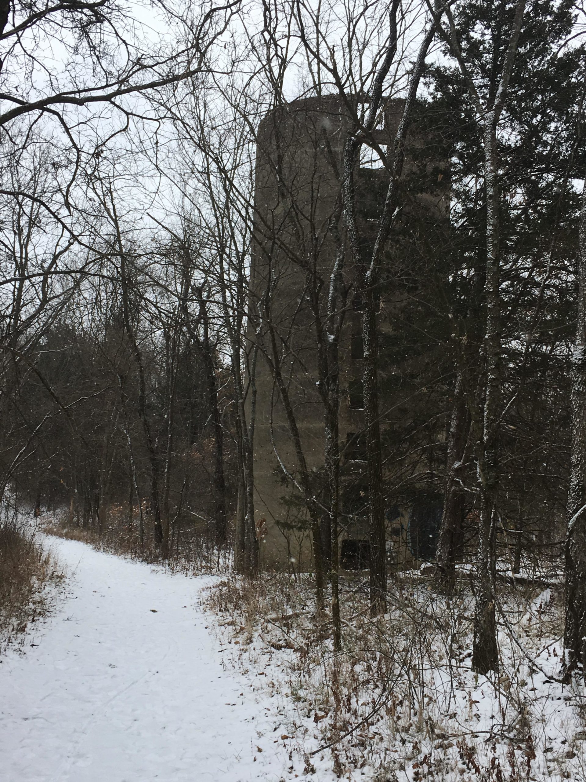A snowy path winds through a forest, with bare trees on either side. In the background, an old, weathered structure, partially obscured by foliage, looms amid the wintry landscape. Rock Bridge Memorial State Park mountain bike trail.