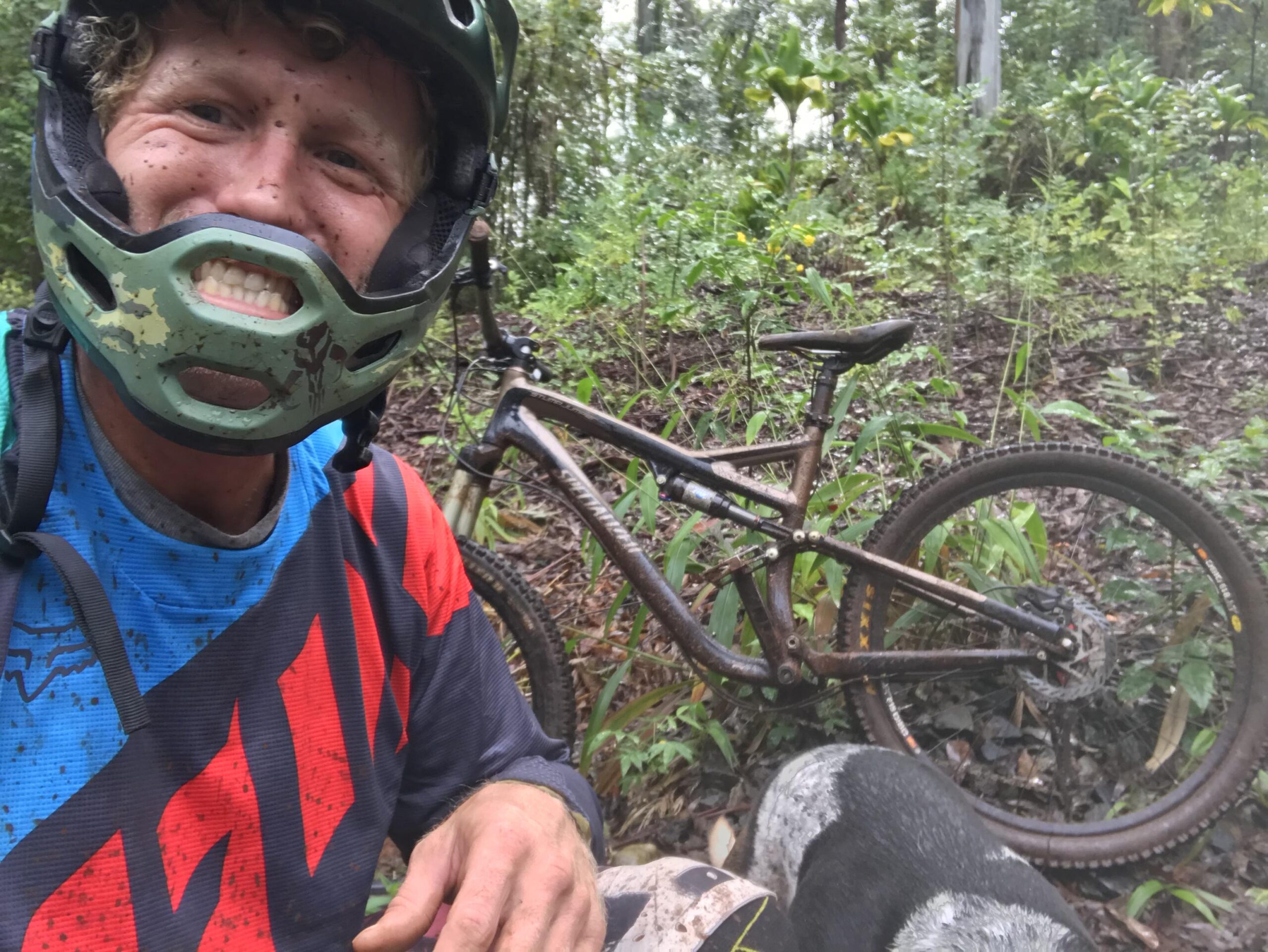 Specialized Stumpjumper FSR: A smiling individual wearing a green helmet and a colorful cycling jersey is sitting on the ground, surrounded by lush greenery. Their face is splattered with mud, and a muddy mountain bike is positioned nearby. A dog is partially visible in the foreground.