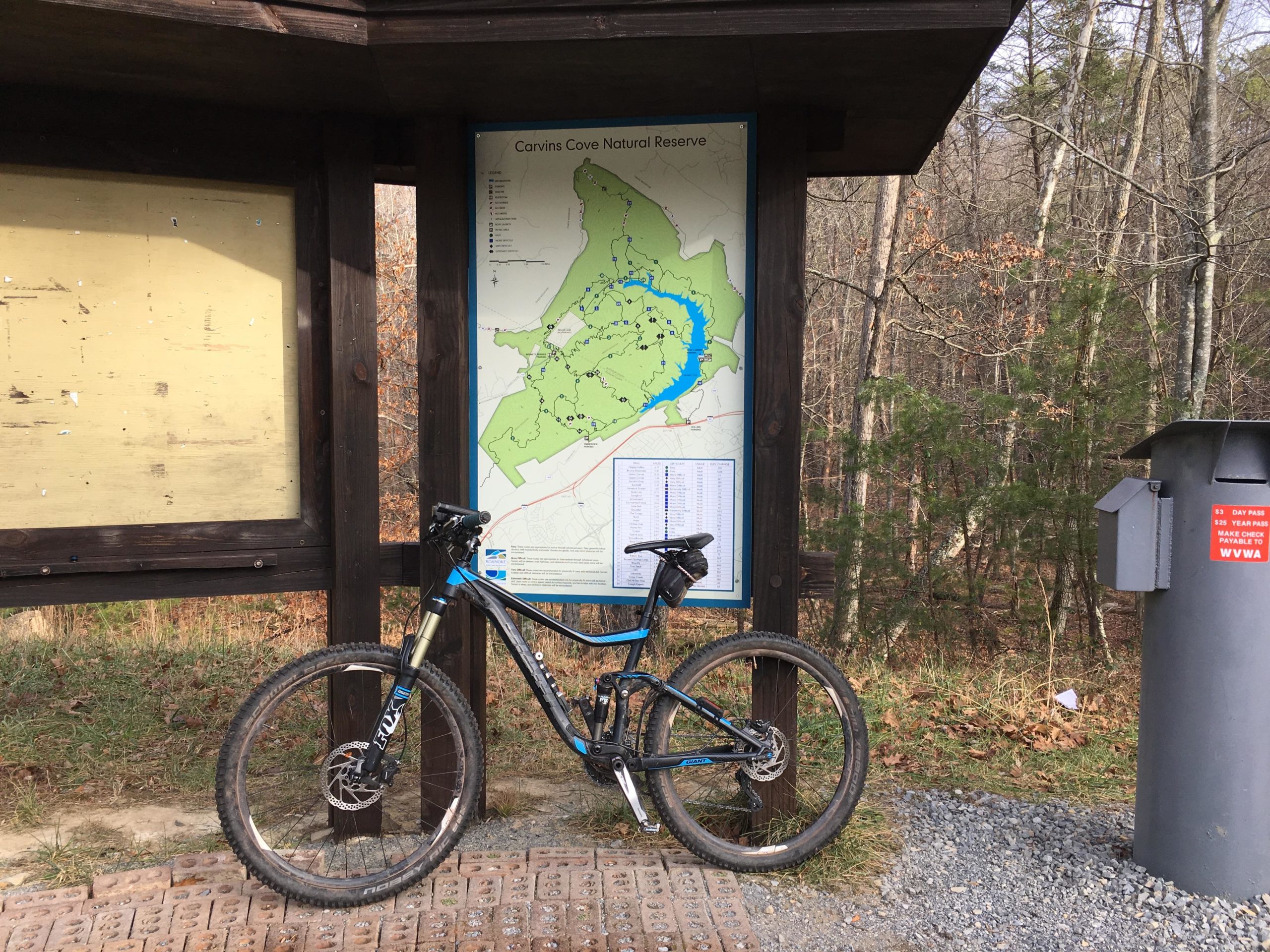 A mountain bike leaning against a map sign at the entrance of Carvins Cove Natural Reserve, surrounded by trees and gravel pathways. The map displays trails and features of the reserve, with a nearby informational kiosk and a fee collection box. Carvin's Cove Trail system mountain bike trail.
