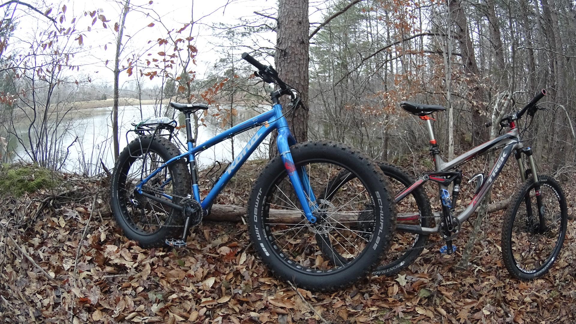 Two mountain bikes, one blue and one gray, are parked next to a tree in a forested area. The ground is covered with fallen leaves, and a calm body of water is visible in the background. The scene conveys a tranquil outdoor setting suitable for cycling. Farris Park mountain bike trail.