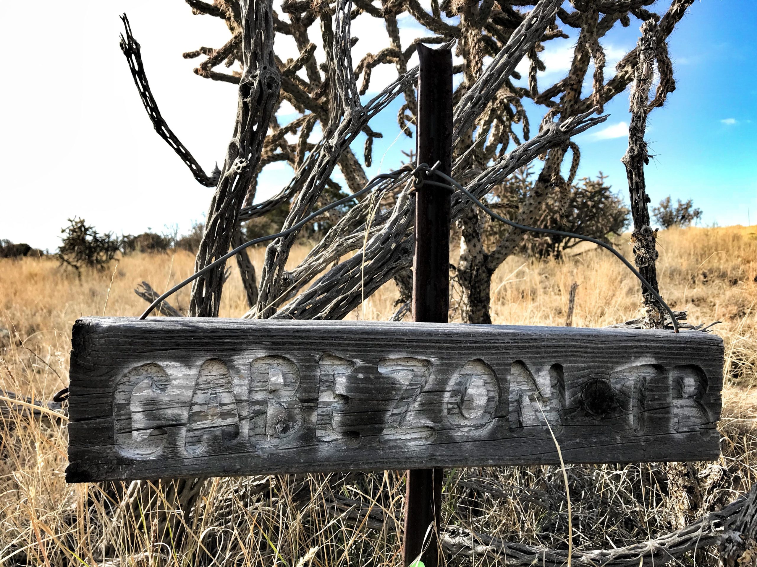 Wooden trail sign that reads "Cabezon TR," positioned in a grassy, arid landscape with sparse vegetation and a clear blue sky in the background. Mariposa Fat Bike Trails mountain bike trail.