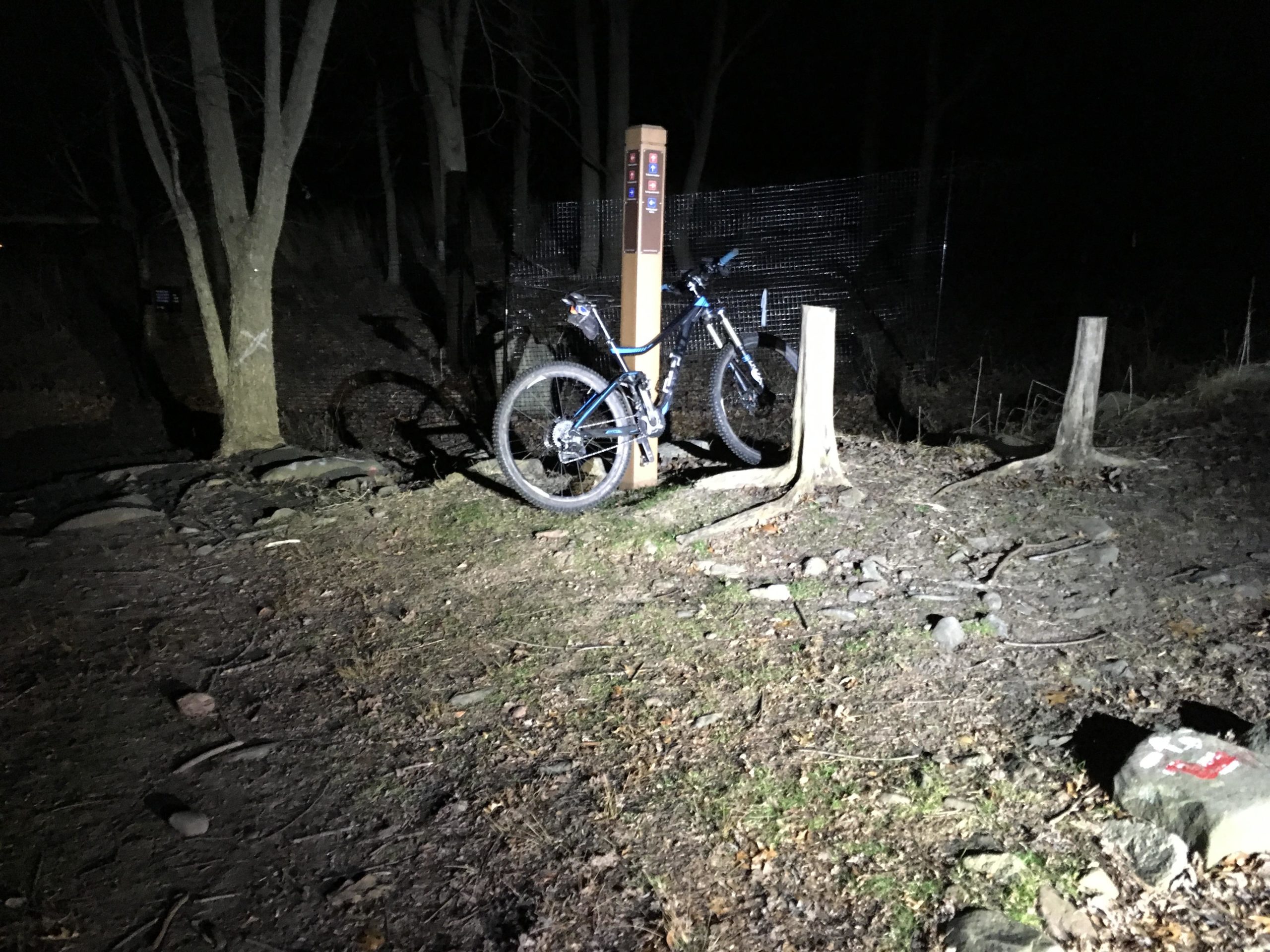 A mountain bike parked near a trail sign in a dark wooded area, illuminated by a flashlight. The surroundings include sparse trees, dirt, rocks, and fallen branches, with faint light highlighting the trail's features. Richmond Avenue and Forest Hill road mountain bike trail.
