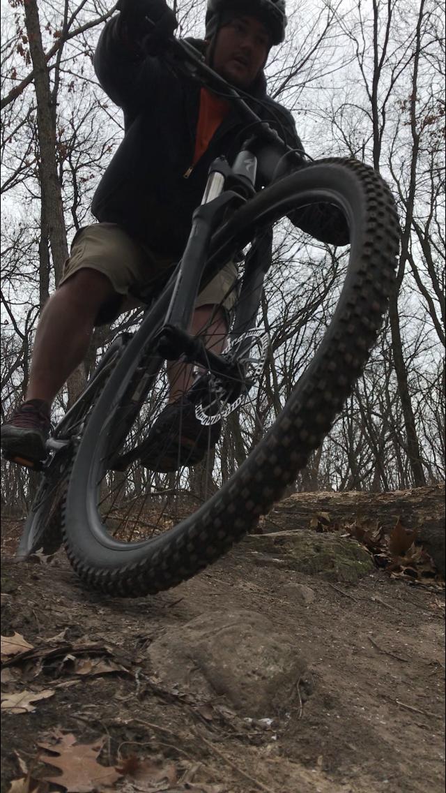 A person riding a mountain bike on a rugged trail, leaning forward as they navigate over a rock, surrounded by trees in a winter landscape. The focus is on the front wheel of the bike and the rider's determined expression. Kettle Moraine John Muir + Emma Carlin mountain bike trail.